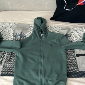 Grön hoodie från US Polo Assn - Säljer en grön hoodie från US Polo Assn med dragkedja och huva. Tröjan har en broderad logga på bröstet och är perfekt för en avslappnad stil. Passar bra till både jeans och joggers.
