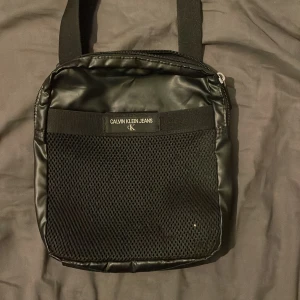  Calvin Klein väska  - Snygg svart axelväska från Calvin Klein Jeans med ett stilrent nätfack på framsidan och dragkedja upptill. Perfekt för att bära med sig det viktigaste. Väskan har justerbar axelrem för bekväm passform.