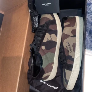 Camouflage sneakers från Saint Laurent - Snygga sneakers från Saint Laurent i ett coolt camouflage-mönster. Skorna har en klassisk design med snörning och en bekväm sula. Perfekta för att ge din outfit en edgy touch.