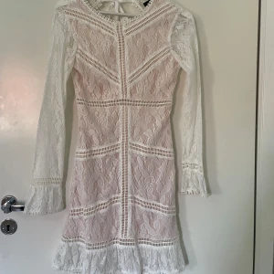 Vit spetsklänning med öppen rygg - Elegant vit spetsklänning med långärmad design och öppen rygg. Klänningen har vackra spetsdetaljer och volanger vid ärmslut och nederkant. Perfekt för speciella tillfällen, som studenten som snart närmar sig!