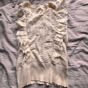 Beige spetsprydd topp från Lindex - Säljer en söt beige topp från Lindex  med spetsdetaljer och volanger längs sidorna. Toppen har en ribbad nederkant och ett vackert mönster som ger en romantisk känsla. Perfekt för att piffa upp garderoben!