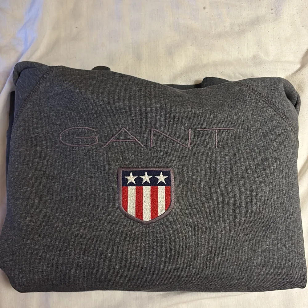 Säljer en stilren grå hoodie från GANT med broderad logga och emblem på bröstet. Tröjan har en klassisk design med huva och långa ärmar, perfekt för en avslappnad look. Den är mjuk och bekväm, idealisk för kyligare dagar.. Neuletakit & Villapaidat.