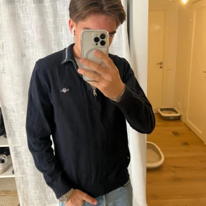 Svart tröja med dragkedja från GANT - Säljer en stilren svart tröja från GANT med dragkedja vid halsen. Tröjan har långa ärmar och ett diskret broderat märke på bröstet. Perfekt för en avslappnad och snygg look.