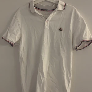 Vit kortärmad pikétröja från Moncler - Säljer en stilren vit pikétröja från Moncler med korta ärmar och klassisk krage. Tröjan har en broderad logotyp på bröstet och röd-svarta detaljer på krage och ärmslut. Perfekt för en avslappnad men snygg look. Strl: S passar M (för att få referens bild på hur den ser ut på mig skriv privat) OBS: en defekt på axeln (se bild) men inget som syns när man har tröjan på sig! pris går att diskutera 