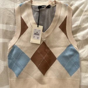 Beige väst med rutor från BikBok - Säljer en beige stickad väst från BikBok med klassiskt rutig argyle-mönster i brunt och ljusblått. Västen har v-ringning och ribbade kanter, perfekt att styla över en skjorta eller t-shirt.