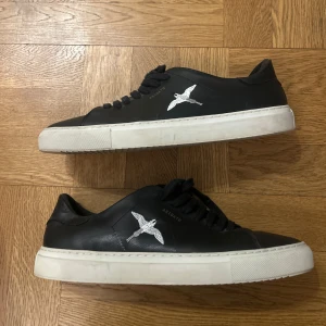 Svarta sneakers från Axel Arigato - Säljer ett par svarta sneakers från Axel Arigato med vit sula och broderad fågel på sidan. Skorna har snörning och rund tå, tillverkade i skinn. Perfekta för en stilren och modern look. Har tyvärr inte kvar lådan!