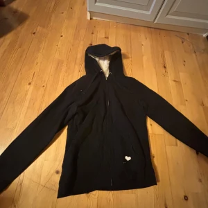 Svart hoodiejacka med fuskpälsfoder från Fishbone - Mysig svart hoodiejacka från Fishbone med dragkedja och huva. Insidan är fodrad med mjuk, vit fuskpäls. Jackan har ett litet hjärta framtill och texten '...love it' baktill. Perfekt för kyliga dagar.