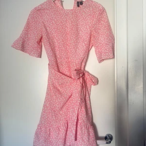 Rosa blommig omlottklänning - Söt rosa omlottklänning med vitt blommönster och knyt i midjan. Klänningen har korta ärmar och volang nertill, perfekt för varma dagar. Helt oanvänd! 💓