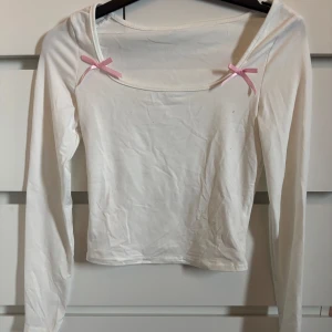 Vit långärmad topp med rosetter - Söt vit långärmad topp med två rosa rosetter vid axlarna och en bred, fyrkantig urringning. Perfekt för en gullig och stilren look.