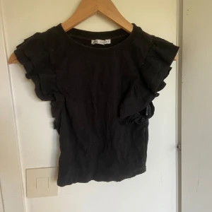 Svart volangtopp från Zara - Svart topp från Zara med volangdetaljer längs ärmarna. Tillverkad i mjuk bomull med 5% elastan för extra stretch. Kortärmad och croppad modell som ger en trendig look.