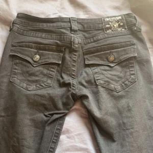 Svarta jeansbyxor från True Religion - Säljer ett par svarta jeans från True Religion med klassiska bakfickor och dekorativa sömmar. Byxorna har knappar på fickorna och en tydlig logga på bakre linningen. Storlek W27 men skulle säga W28, längd 32