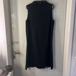 Klänning  - Säljer en svart ärmlös klänning med hög hals och slits framtill. Klänningen har en rak och enkel design som ger en stilren look. Perfekt för dig som gillar minimalistiskt mode.