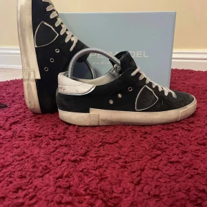 Svarta Philippe Model - Säljer ett par svarta Philippe Model sneakers med vita sulor och vita snören. Skorna har en ikonisk sköld-detalj på sidan och silverfärgade öljetter. Klassisk låg modell med rund tå och platt sula. Perfekta för en stilren vardagslook.