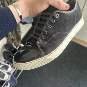 Svarta sneakers från Lanvin med lackad tå - Säljer ett par svarta sneakers från Lanvin med ovandel i mocka och en glansig, lackad tå. Skorna har vita sulor och klassisk snörning. Perfekta för dig som vill ha en stilren men ändå unik sneaker.