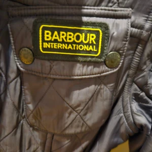 Svart quiltad jacka från Barbour International - Säljer en svart/lila quiltad jacka från Barbour International med flera fickor och knappar framtill. Jackan har hög krage, bälte i midjan och klassisk Barbour-logga på bröstet. Perfekt för dig som gillar tidlös och sportig stil.