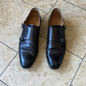 Svarta skinnloafers med dubbla spännen - Stilrena svarta loafers i skinn med dubbla spännen i metall. Skorna har fyrkantig tå och låg platt sula, perfekt för en dressad look. Insidan är fodrad i ljust skinn och de har en elegant, minimalistisk design.