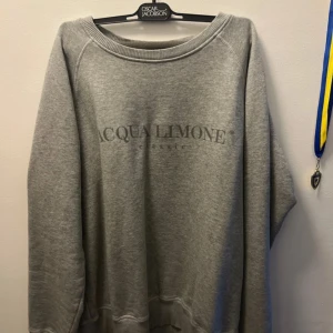 Grå sweatshirt från Acqua Limone - Grå sweatshirt från Acqua Limone med tryckt logga framtill och stort citrontryck i gult på ryggen. Tröjan har rund halsringning och långa ärmar. Aldrig använd men utan prislapp, originalpris: 1099kr