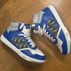 Blå och vita Dior B27 sneakers - Säljer ett par höga sneakers från Dior i blått och vitt med grå detaljer. Skorna har snörning, rund tå och den klassiska Dior-loggan på sidan i ett mönstrat tyg. Perfekta för dig som vill sticka ut med exklusiva sneakers.