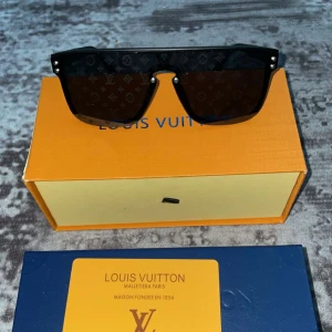 Svarta solglasögon från Louis Vuitton - Stilrena svarta solglasögon från Louis Vuitton med monogrammönster på glasen. Rektangulär, oversized modell med bred båge och diskreta metalldetaljer. Perfekt för dig som vill sticka ut med en lyxig accessoar.