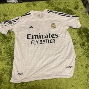 Real Madrid fotbollströja Mbappé 9 - Säljer en vit Real Madrid fotbollströja från Adidas med svarta detaljer och tryck. Tröjan har korta ärmar, klubbmärke på bröstet, sponsortryck och Mbappé 9 på ryggen. Champions League-märke på ärmen och HEAT.RDY-material för extra komfort.