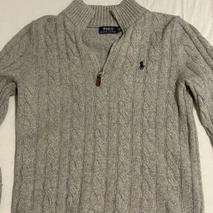 Grå kabelstickad tröja från Polo Ralph Lauren - Snygg grå kabelstickad tröja från Polo Ralph Lauren med halv dragkedja och broderad logga på bröstet. Tröjan har hög krage och långa ärmar, perfekt för lager-på-lager-stil. Pris går att diskutera