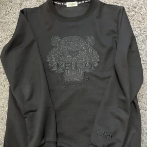 Svart sweatshirt med broderad tiger från Kenzo - Säljer en svart sweatshirt från Kenzo med ikonisk broderad tiger och Kenzo Paris-logga på bröstet. Tröjan har rund halsringning och ribbade muddar. Perfekt för dig som gillar stilrena och trendiga plagg.