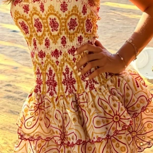 Rosa klänning - Superfin kortklänning med smockad överdel och volangkjol. Klänningen har ett färgglatt mönster i rött och gult på vit botten, med blommiga och geometriska detaljer. Ärmlös modell som passar perfekt till sommarens alla tillfällen.