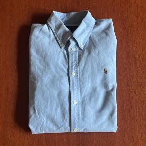 Ljusblå skjorta från Ralph Lauren - Klassisk ljusblå skjorta från Ralph Lauren med button-down krage och broderad logga på bröstet. Skjortan har långa ärmar och vita knappar framtill. Perfekt för en stilren och tidlös look. Har en fläck långt ner på magen och längst ner på armen. storlek XL men passar mer L tror jag.