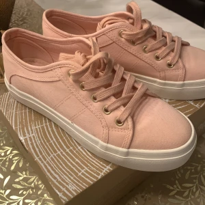 Ljusrosa sneakers med vita sulor - Säljer ett par ljusrosa sneakers från North alaska Riley med klassisk design. Skorna har vita, platta gummisulor och snörning med guldfärgade öljetter. Tillverkade i textilmaterial och passar perfekt till en avslappnad stil.