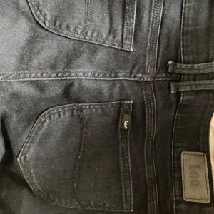 Svarta jeansbyxor från Lee - Säljer ett par svarta jeans från Lee med klassisk femficksdesign och normal passform. Byxorna har bälteshällor och Lee-logga på bakfickan samt läderpatch i midjan. Perfekta till många olika outfits.