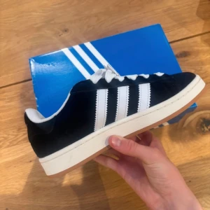 Svarta Adidas Campus sneakers med vita ränder - Klassiska Adidas Campus sneakers i svart mocka med vita ränder och vit sula. Skon har snörning och en gummisula med bra grepp. Perfekt för dig som gillar en stilren och tidlös look.