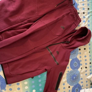 Vinröd hoodiejacka från Nike - Säljer en vinröd hoodiejacka från Nike med hel dragkedja och huva tillsammans med byxa. Jackan har svarta detaljer vid dragkedjorna och en sportig look. Perfekt för dig som gillar streetwear och vill ha något bekvämt och snyggt. Snörena är lite sönder men går att fixa nya i affär. Byxan är i storlek M och hoodiejackan är i storlem S.