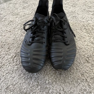 Svarta fotbollsskor från Umbro - Säljer ett par svarta fotbollsskor från Umbro med snörning och mönstrad ovandel. Skorna har rund tå och fasta dobbar för spel på gräs. Klassisk design med Umbro-logga på sidan och platt sula för bra grepp på planen.