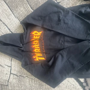 Svart Thrasher hoodie med flames-logga - Svart hoodie från Thrasher med den klassiska flames-loggan i gult och rött på bröstet. Tröjan har huva med dragsko och en stor magficka. Perfekt för dig som gillar streetwear och skatekultur.