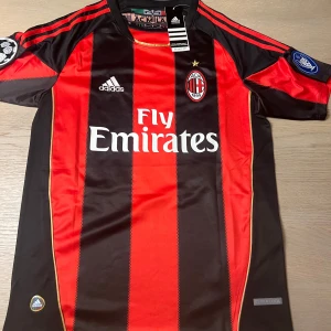 AC Milan fotbollströja Ronaldinho 80 - Säljer en klassisk AC Milan fotbollströja med Ronaldinho och nummer 80 på ryggen. Tröjan är svart och röd med vertikala ränder, korta ärmar och Adidas-logga samt Fly Emirates sponsortryck på bröstet. Klubbmärke på vänster bröst och Champions League-märke på ärmen.