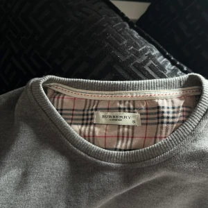 Grå tröja från Burberry - Säljer en klassisk grå tröja från Burberry med rund halsringning och det ikoniska rutiga mönstret på insidan av kragen. Tröjan har långa ärmar och en liten broderad Burberry-logga på bröstet.