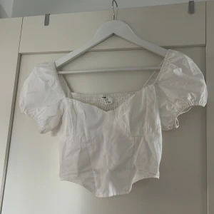 Vit croppad blus med puffärmar - Säljer en söt vit croppad blus med puffiga korta ärmar och smockad rygg. Toppen har en fyrkantig ringning och är perfekt för en trendig look. 