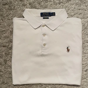 Ralph Lauren Piké  - Storlek L men sitter mer som S/M .Klassisk vit pikétröja med korta ärmar från Polo Ralph Lauren med broderad logga på bröstet. Tröjan har krage, tre knappar framtill och är tillverkad i bomull. Perfekt för en stilren och tidlös look.