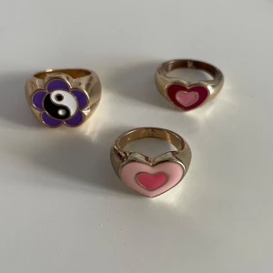 Tre chunky ringar med hjärtan och blomma - Säljer tre coola ringar i guldtonad metall. Två av ringarna har hjärtan i rosa och rött, och en ring har en lila blomma. Perfekta för att ge din stil en färgglad och lekfull touch!