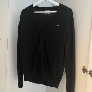 Svart kofta från J.Lindeberg - En klassisk svart kofta från J.Lindeberg med v-ringad hals och diskret logga på bröstet. Koftan har långa ärmar och knäppning framtill. Perfekt för lager-på-lager och enkel att matcha.