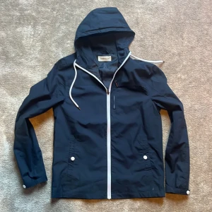 Mörkblå vindjacka från Pier One - Snygg mörkblå vindjacka från Pier One med vit dragkedja och huva med dragsko. Jackan har två fickor framtill med vita knappar och en stilren, enkel design. Perfekt för blåsiga dagar och enkel att matcha med olika outfits.