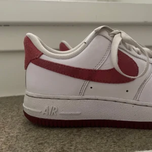 Nike Air Force 1  - Säljer ett par vita Nike Air Force 1 med röd mocka-swoosh och röd detalj på hälen. Skorna har klassisk låg modell, perforerad tå och vit sula med röd undersida. Perfekta för dig som vill sticka ut lite extra med dina sneakers.