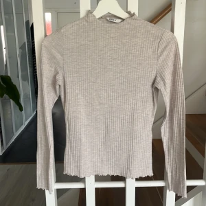 Beige ribbad långärmad topp från ONLY - Säljer en beige ribbad långärmad topp från ONLY. Tröjan har en smal passform och diskret hög hals. Materialet är mjukt och stretchigt, perfekt för lager-på-lager. Avslutet nertill och vid ärmslut har en vågig kant för extra detalj.
