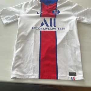 PSG vit fotbollströja med röd och blå rand - Vit PSG fotbollströja med röd och blå vertikal rand, Nike-logga och klubbmärke på bröstet. Tryck med sponsortext på framsidan. Klassisk design med korta ärmar och rund hals.