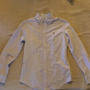 Ljusblå skjorta från Ralph Lauren - Säljer en klassisk ljusblå skjorta från Ralph Lauren med broderad logga på bröstet. Skjortan har lång ärm, knappar framtill och en skräddarsydd passform (custom fit). Perfekt för en stilren look!