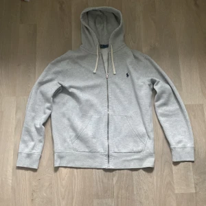 Grå hoodie från Polo Ralph Lauren - Säljer en ljusgrå hoodie från Polo Ralph Lauren med dragkedja och huva. Tröjan har två fickor framtill och klassisk broderad logga på bröstet. Perfekt för en avslappnad stil.
