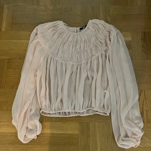 Ljusrosa blus med rynkade detaljer - Säljer en ljusrosa blus med rynkade detaljer upptill och vid ärmslut. Blusen har långa, luftiga ärmar och resår i midjan för en snygg passform. Perfekt för dig som gillar romantisk och feminin stil.