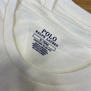 Vit t-shirt från Polo Ralph Lauren - Vit t-shirt från Polo Ralph Lauren med blå broderad logga på bröstet. Klassisk rund hals och korta ärmar. Slim fit-modell i mjuk bomull, perfekt för en clean och stilren look.