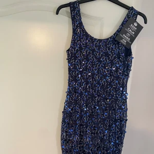 Marinblå glittrig paljettklänning - Säljer en marinblå tight klänning täckt av glittrande paljetter. Klänningen är ärmlös med rund halsringning och har en kroppsnära passform som framhäver siluetten. Perfekt för dig som vill sticka ut på festen! Prislappen är även på.