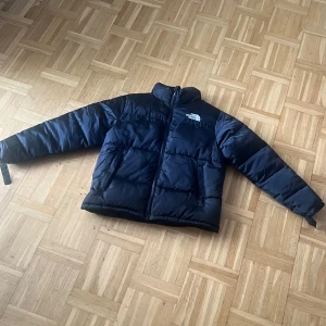 Svart dunjacka från The North Face - Säljer en svart dunjacka från The North Face med hög krage och dragkedja framtill. Jackan har två stora fickor på sidorna och klassisk logga på bröstet och ryggen. Perfekt för kyliga dagar och riktigt skön att bära.